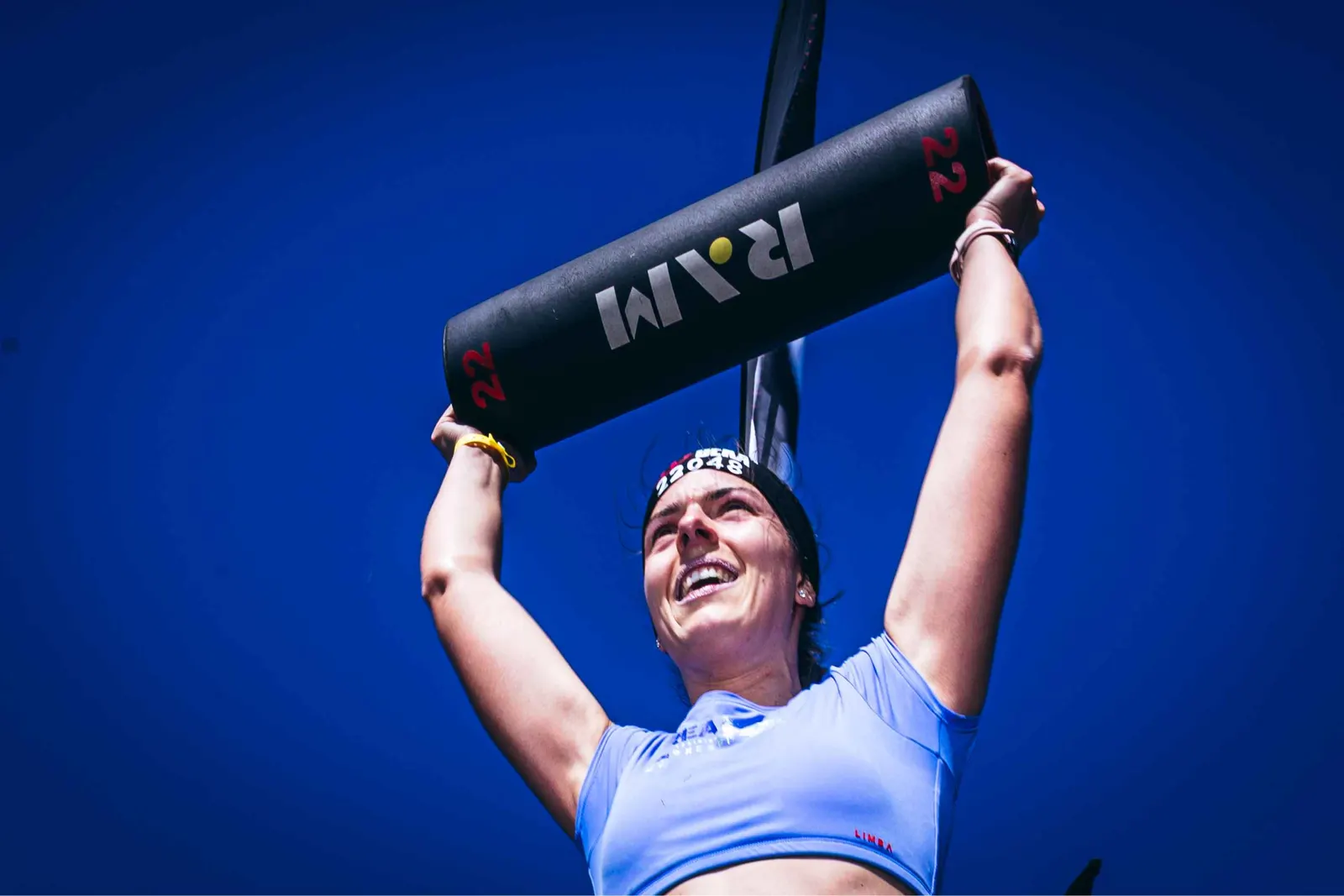 Atleta levantando un saco por encima de la cabeza bajo un cielo azul despejado en una prueba DEKA