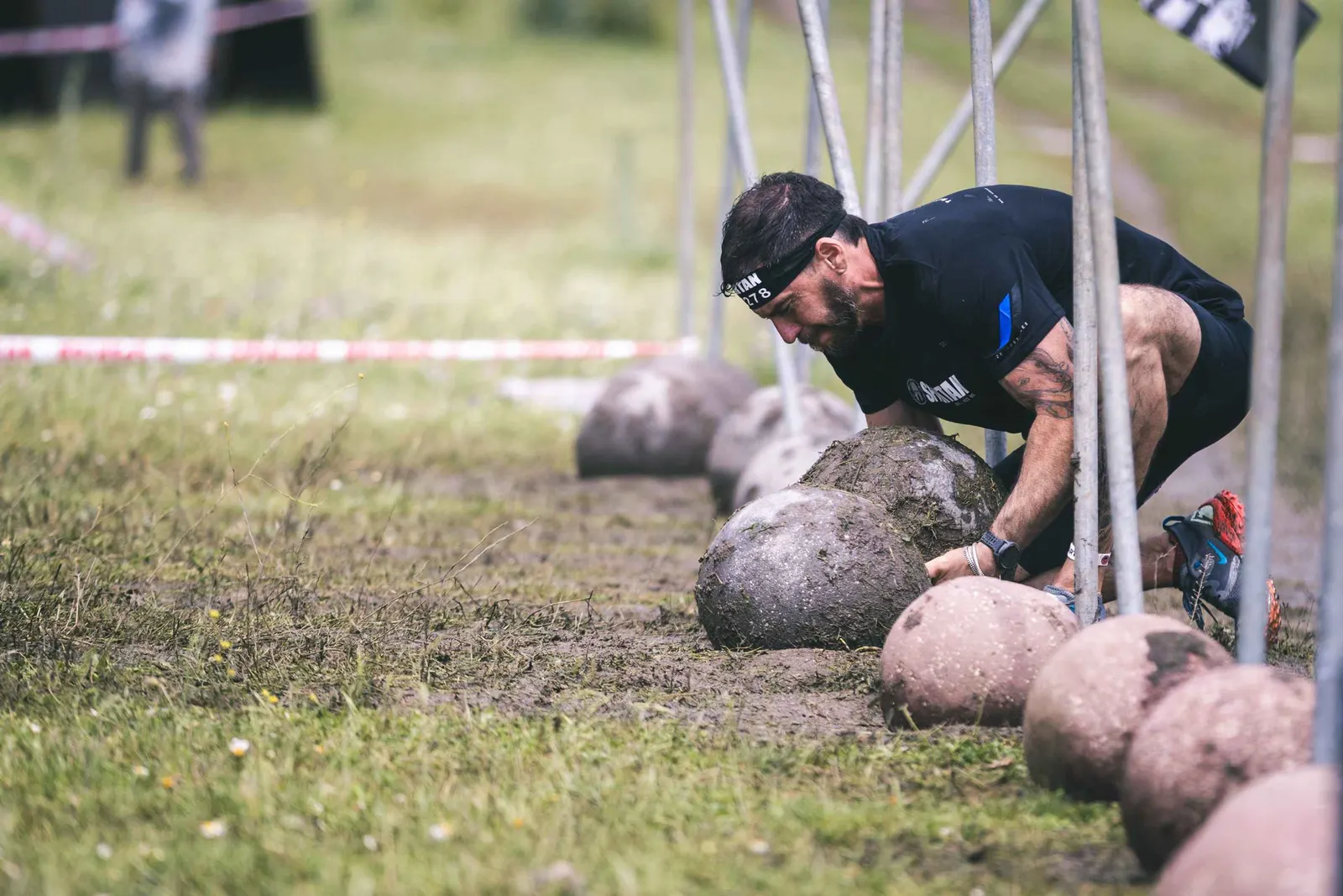 Corredor con cinta Spartan agachado para levantar una bola de piedra en un obstáculo sobre hierba