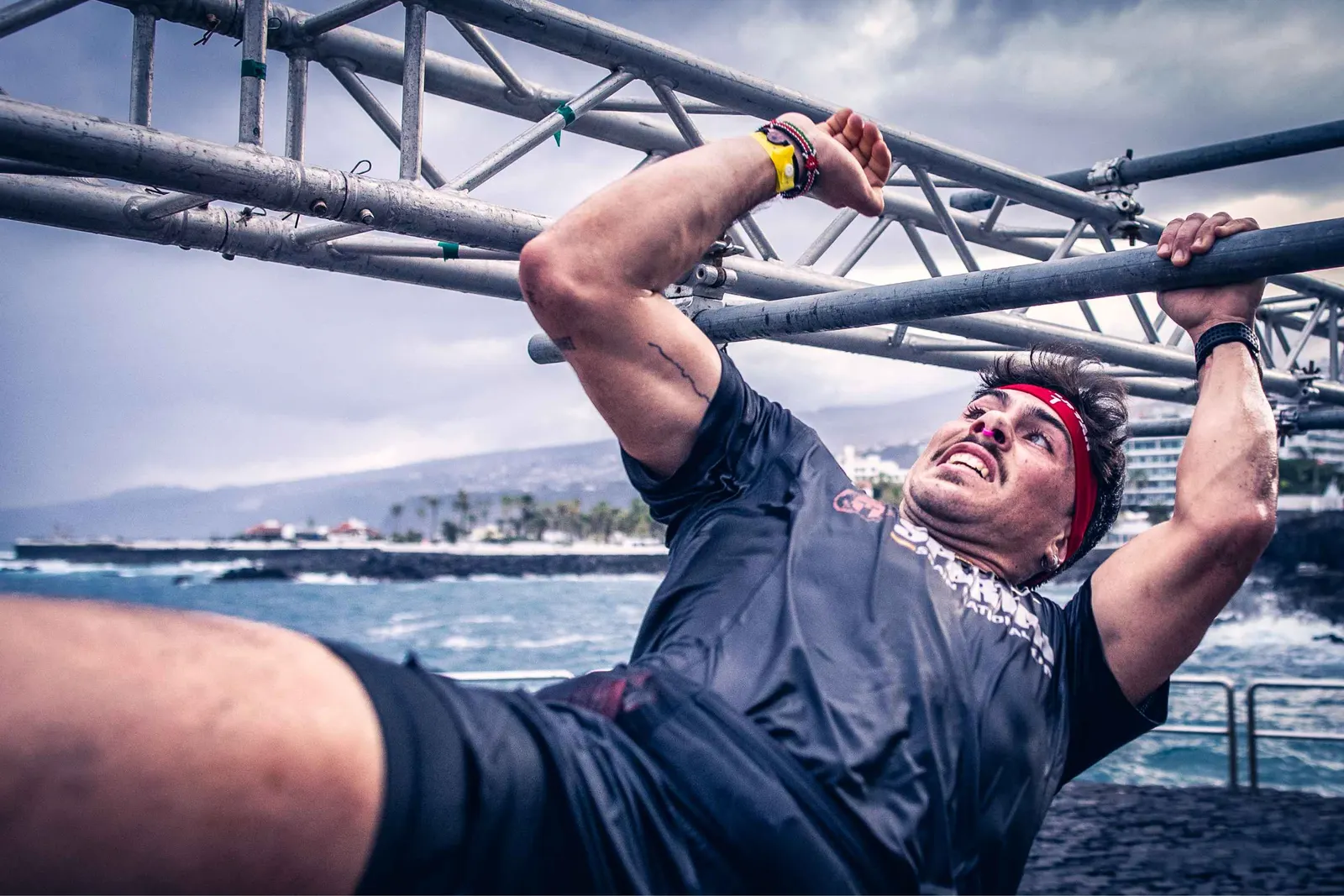 Atleta colgado de una estructura metálica en un obstáculo Spartan junto al mar, con el cielo nublado